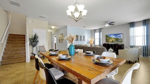 A dining area with a wooden table set for four, surrounded by white chairs. A chandelier hangs above the table, and a television is mounted on the wall to the right. - Unwind in Style: Spacious Villa with Game Room and Private Pool at ChampionsGate (Four Corners)