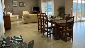 A spacious living area with a sofa, a television, and a dining table with chairs. There is a balcony with a view of the outdoors.