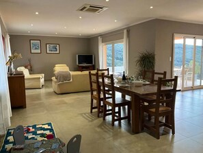 A spacious living area with a sofa, a television, and a dining table with chairs. There is a balcony with a view of the outdoors.