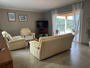 Uma sala de estar com sofá, duas poltronas, mesa de centro, televisão e uma varanda com vista.