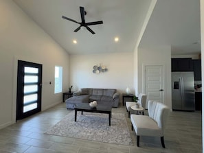 A spacious living room with a sectional sofa, a coffee table, a white armchair, a ceiling fan, and a kitchen area in the background.