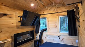 A wooden cabin interior with a bed, a mounted TV, a fireplace, and windows. - Wood Duck Cabin, No Neighbours, Drive too, Pet Friendly (Golden Lake)