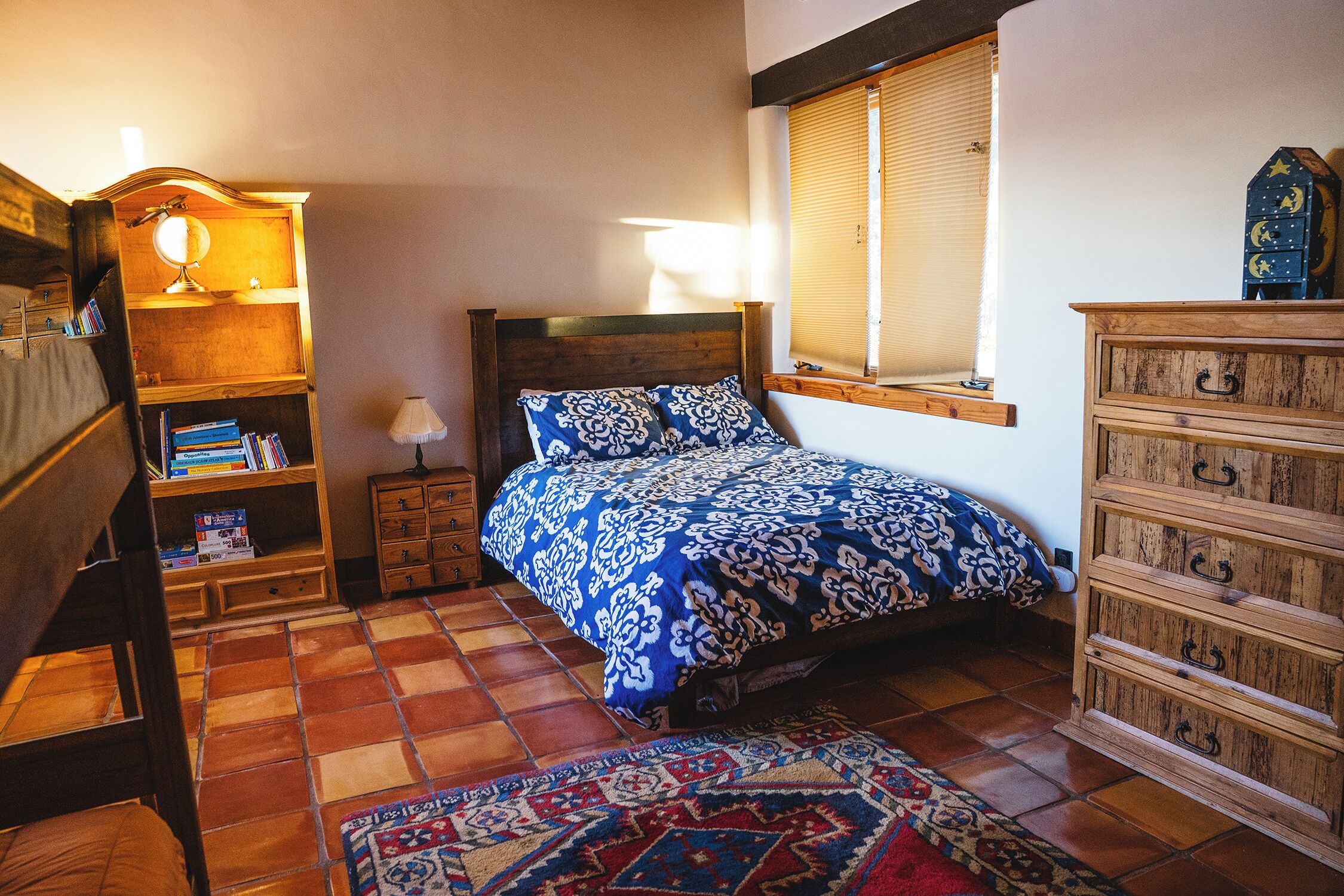 Ein Schlafzimmer mit Bett, einem Holzkleiderschrank, einem Nachttisch mit Lampe, einem Bücherregal und einem Fenster mit Jalousien.