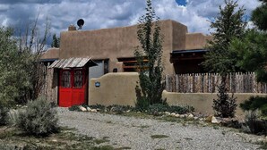 Un bâtiment traditionnel en adobe, avec une porte rouge, entouré de végétation désertique et délimité par une clôture en bois.