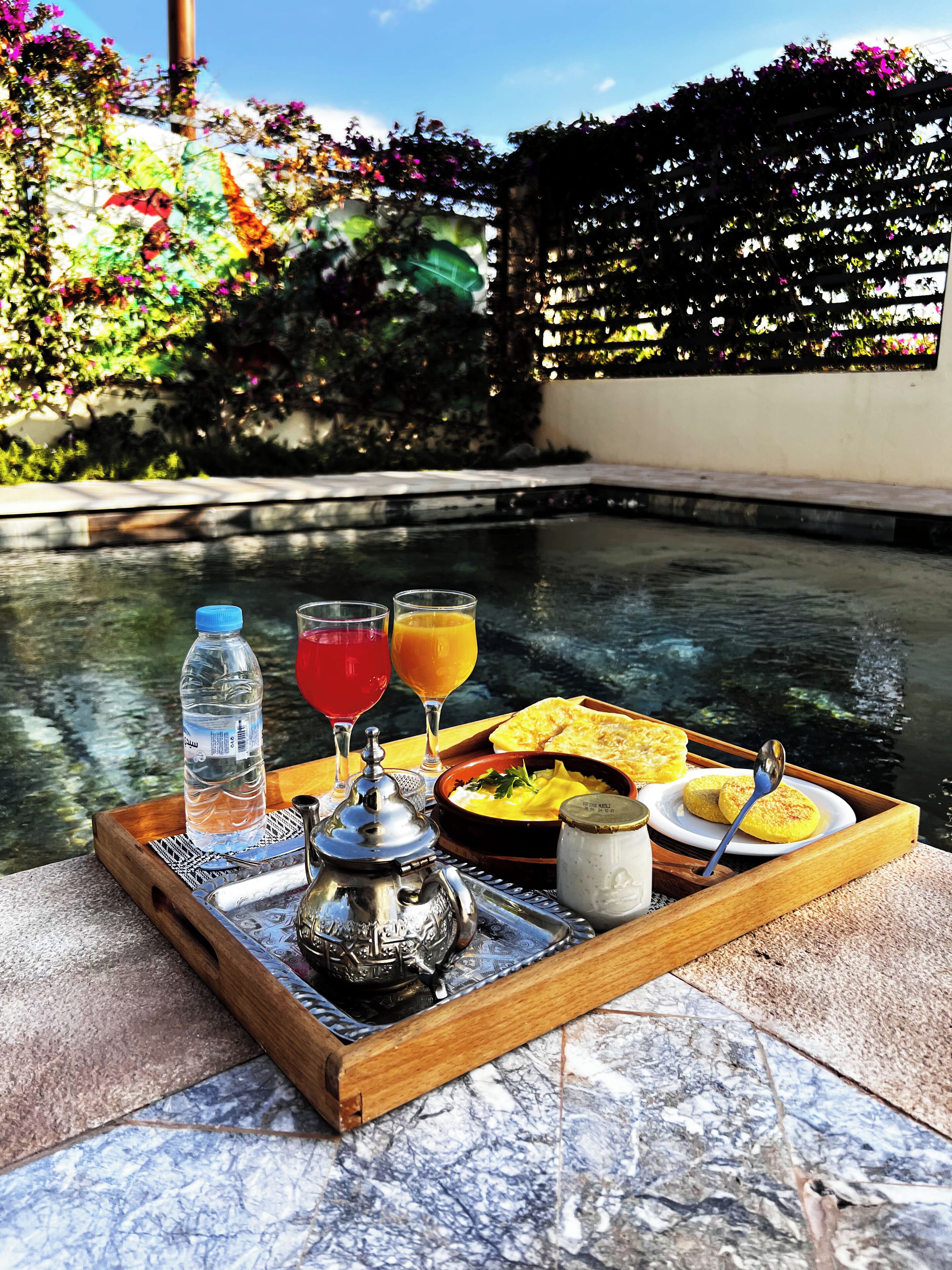 A tray with a teapot, water bottle, glasses of red and orange beverages, and a plate with food and a spoon, set by a pool with a garden in the background.