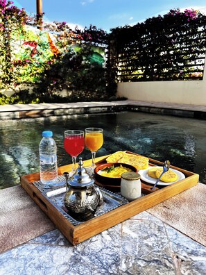 A tray with a teapot, water bottle, glasses of red and orange beverages, and a plate with food and a spoon, set by a pool with a garden in the background. - SOOFSTAY VILLA - Noria Golf Club (Marrakech)