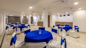 A conference room with round tables, white chairs, and blue tablecloths. There are ceiling fans, recessed lighting, and a wooden cabinet. - The Gleam Hotel (Gurugram)
