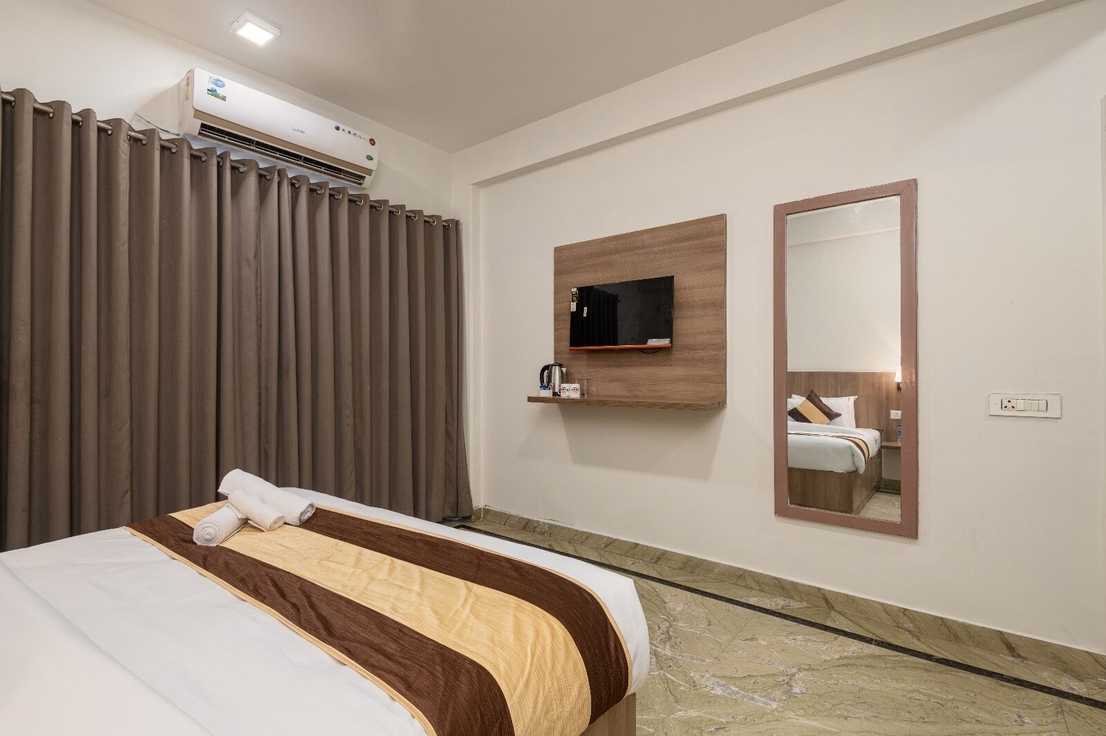 A hotel room with a bed, curtains, an air conditioning unit, a mounted TV, a mirror, and a marble floor.