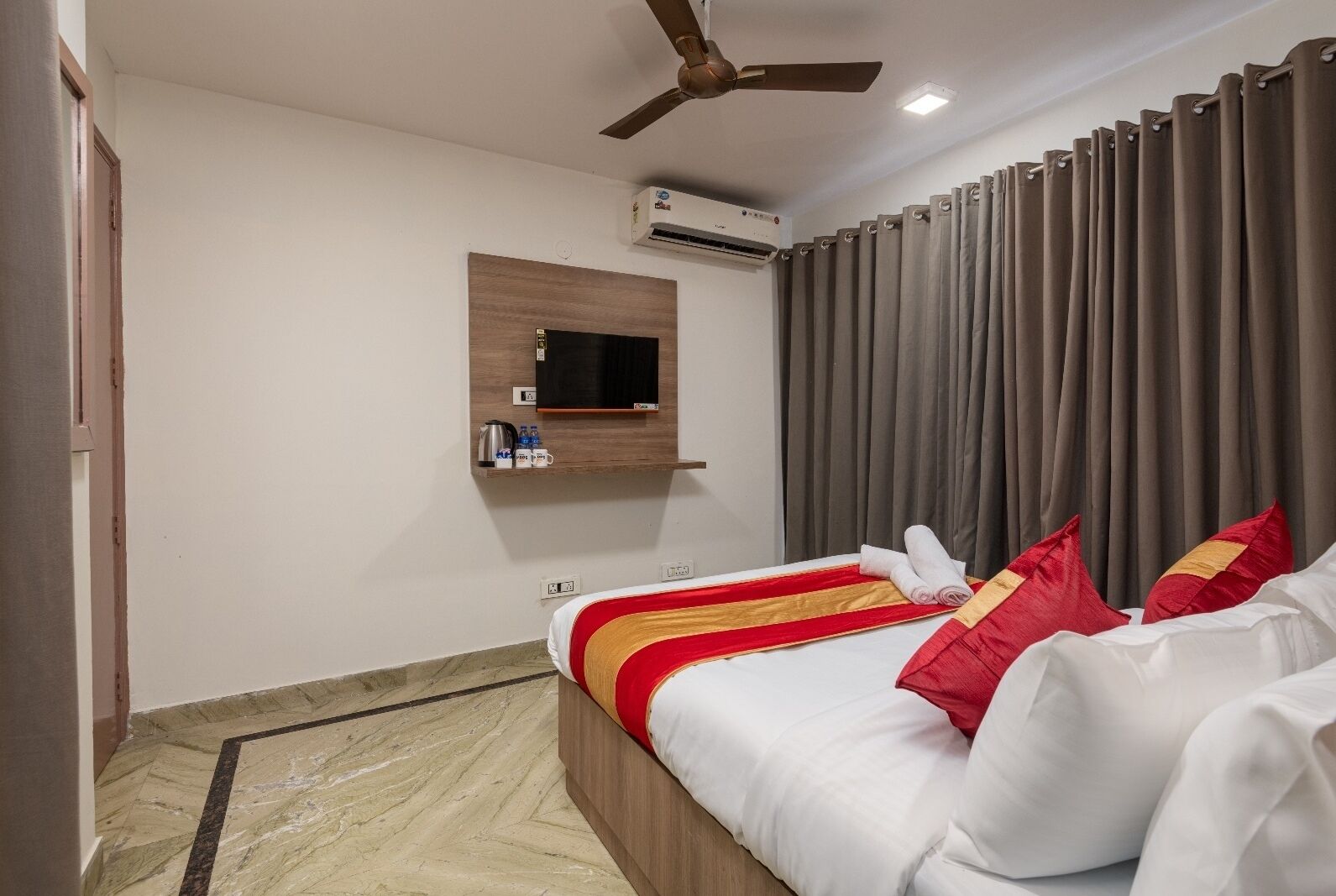 A hotel room with a bed, a ceiling fan, an air conditioning unit, a mounted TV, and a window with curtains.