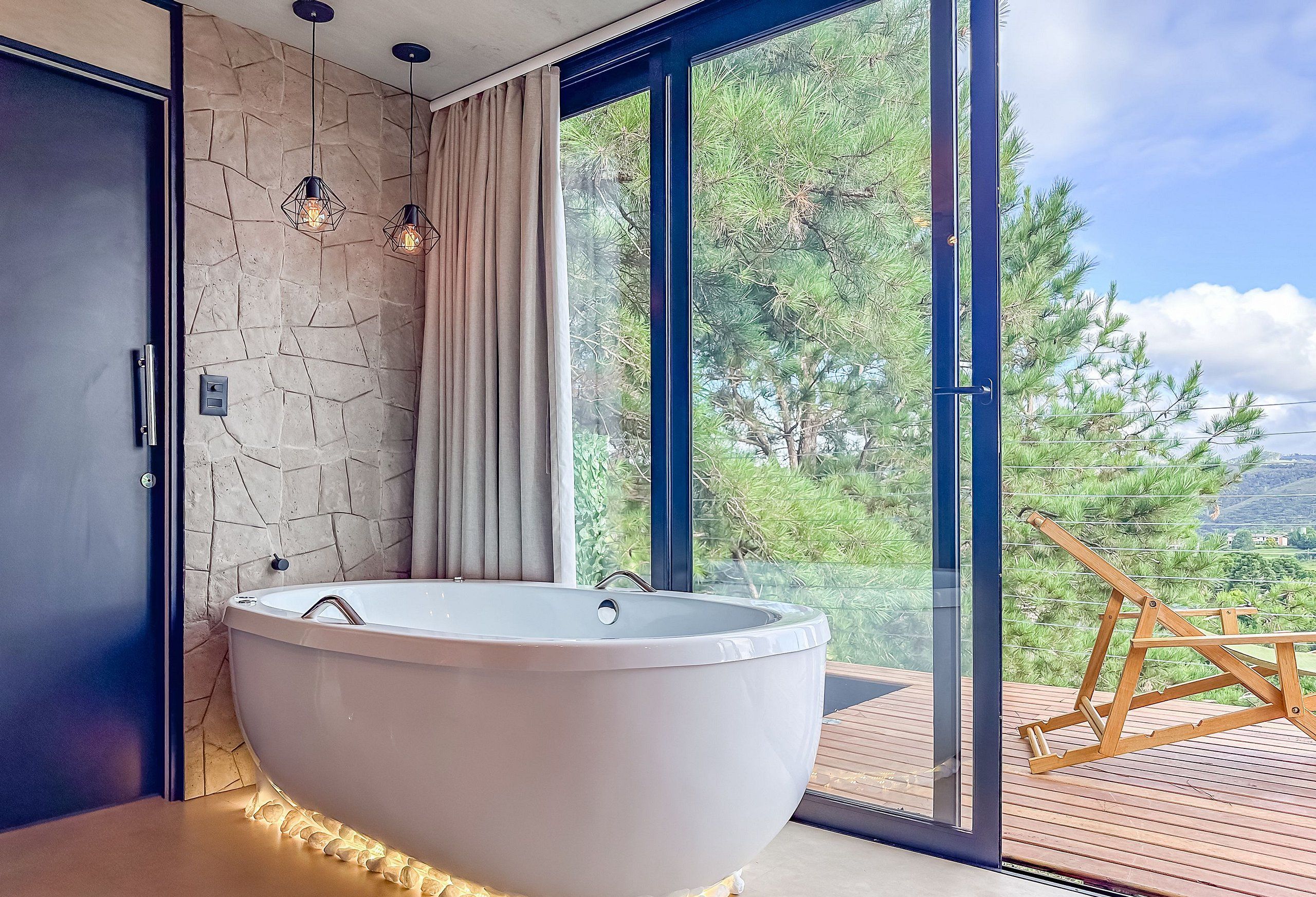 Un baño moderno con una bañera independiente, pared de piedra y un amplio ventanal con vistas a árboles y colinas.