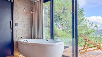 Un baño moderno con una bañera independiente, pared de piedra y un amplio ventanal con vistas a árboles y colinas.