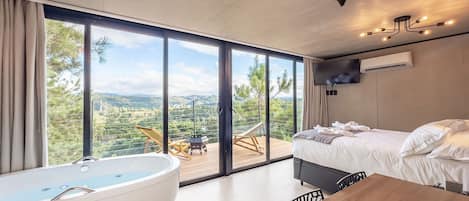 A modern bedroom with a large window, a bathtub, a bed, and a dining area.