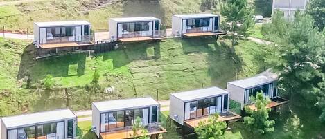 A series of modern, elevated structures with glass facades and wooden decks, situated on a grassy area with trees and a road in the background.
