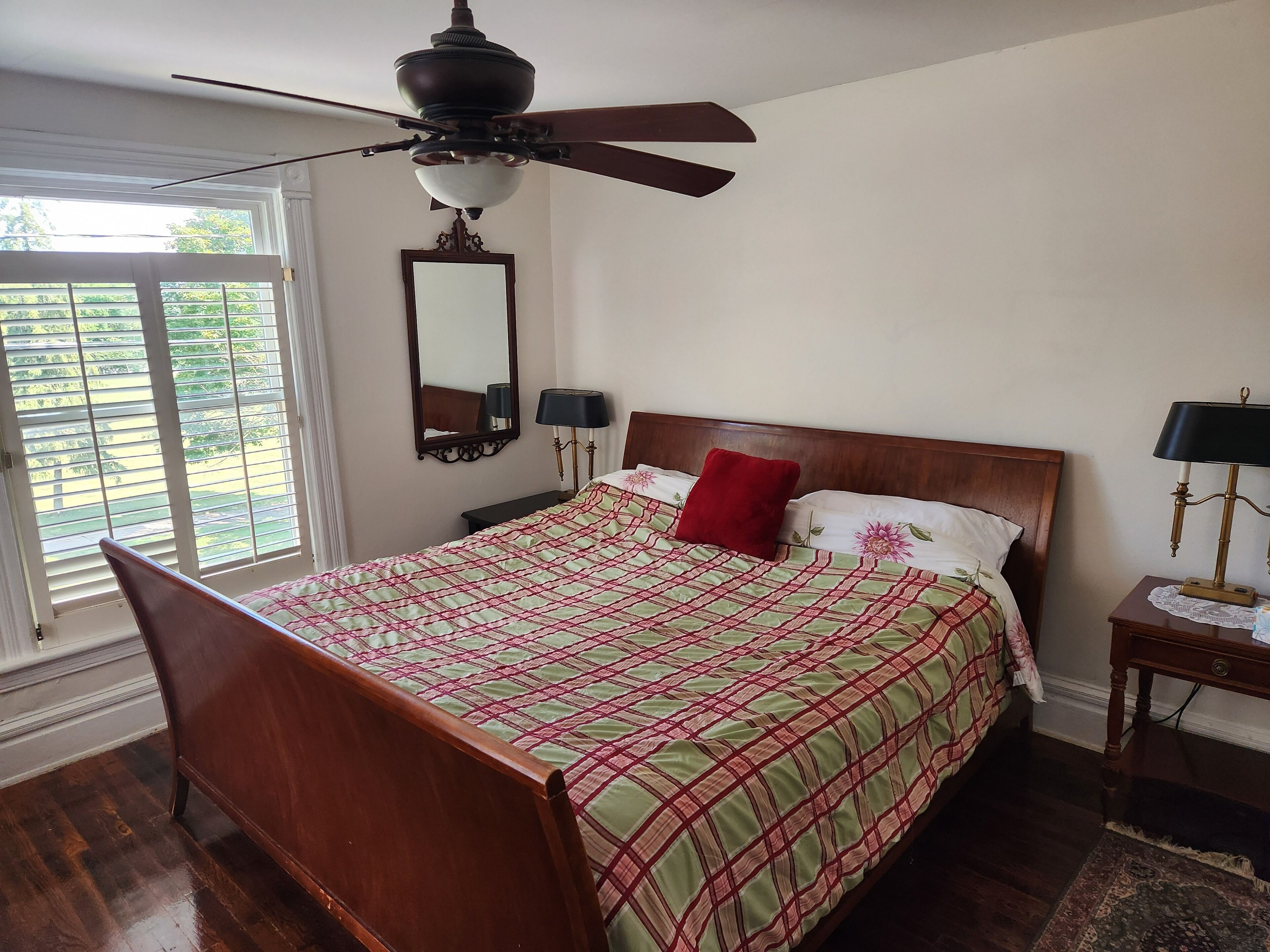 Un dormitorio con cama de madera, ventilador de techo, ventana grande con persianas, mesita de noche con lámpara y un espejo en la pared.