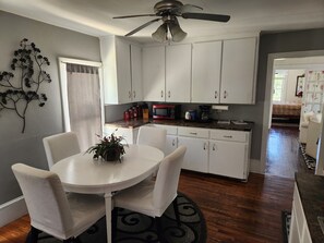 Une cuisine avec des meubles blancs, une table à manger avec des chaises, un ventilateur de plafond et une fenêtre avec un rideau.