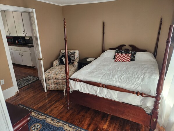 A bedroom with a four-poster bed, a wooden nightstand, a chair with a floral pattern, and a kitchen visible through an open door.