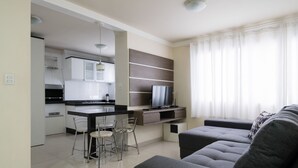 A modern living room with a sofa, a dining area with a bar stool, and a kitchen with white cabinets and a flat-screen TV. - EAC Apto 2min praia Balneário Camboriú (Balneário Camboriú)