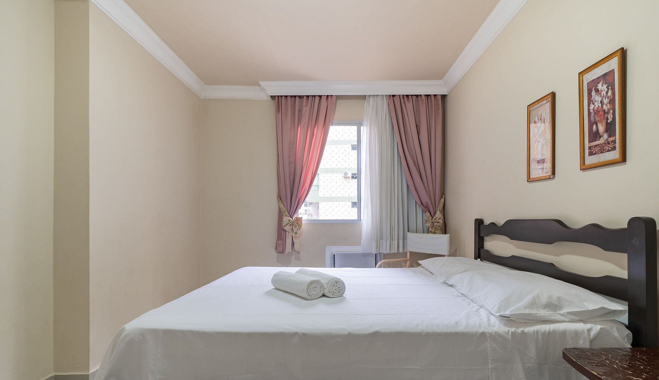 A hotel room with a bed, a nightstand, a window with curtains, and two framed pictures on the wall.