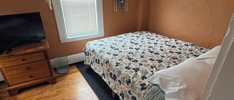 A bedroom with a bed, a dresser, a TV, a window, and a plant.