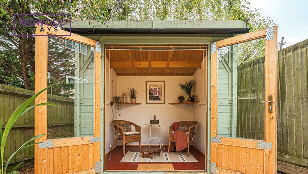 A wooden structure with glass doors, two wicker chairs, a small table, and a framed picture on the wall. - Link Road House By REDWOOD STAYS (Alton)