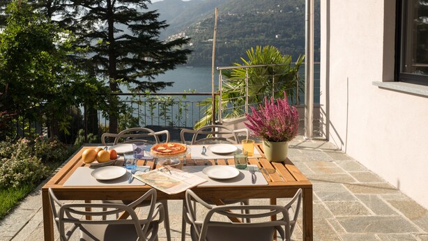 A wooden table set for two with plates, cutlery, and a glass of water on a balcony overlooking a lake and mountains. - Villa Olivo (Blevio)