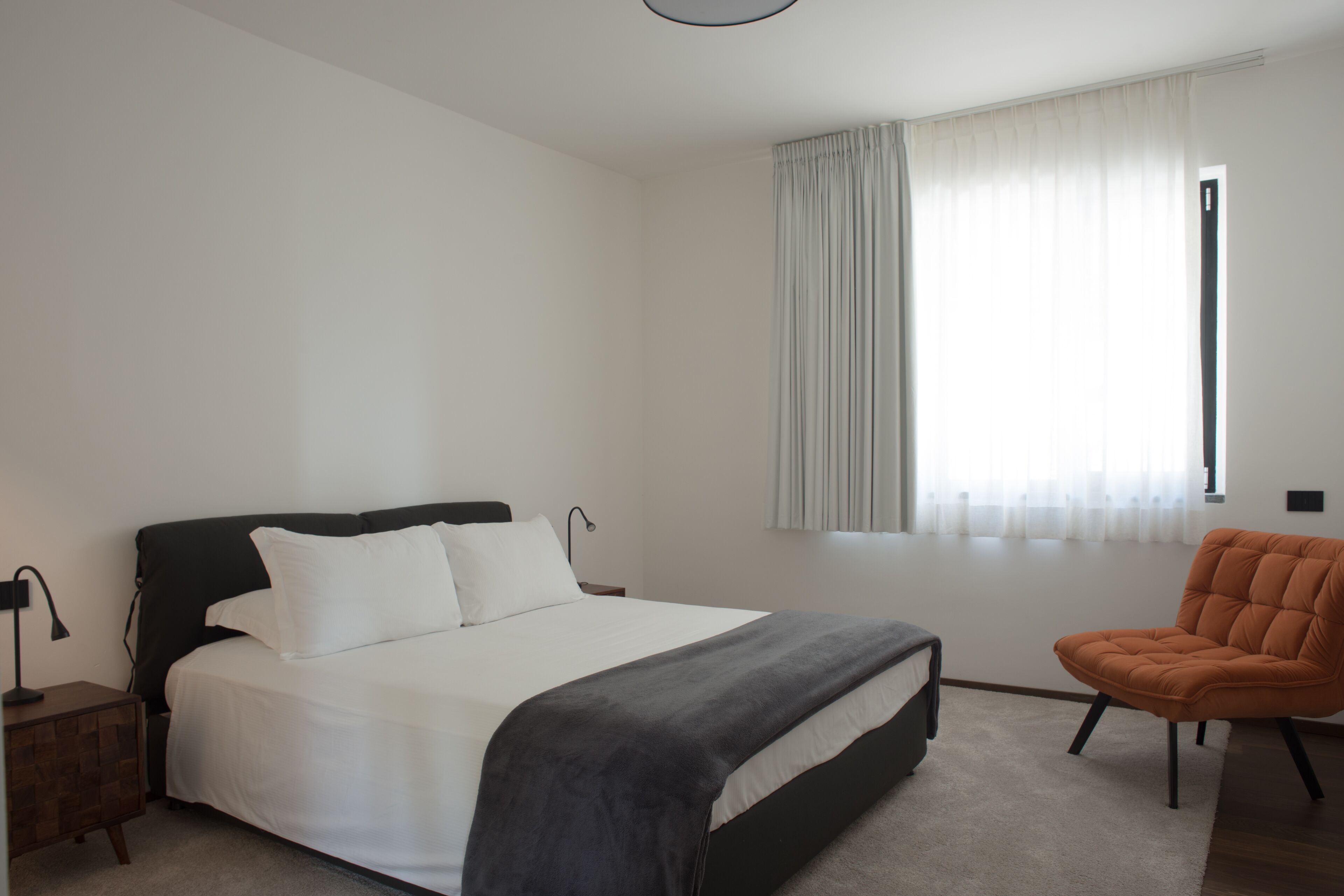 Un dormitorio con cama, una silla, una mesita de noche y una ventana con cortinas.