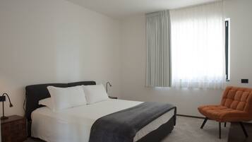 Un dormitorio con cama, una silla, una mesita de noche y una ventana con cortinas.