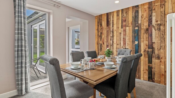 Een eetruimte met een houten tafel gedekt voor zes personen, grijze gestoffeerde stoelen en uitzicht door een glazen deur op een balkon.