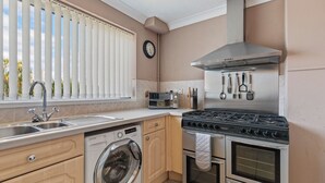 Een kleine keuken met een ingebouwde wasmachine, oven en kookplaat, een spoelbak en een raam met zonwering.
