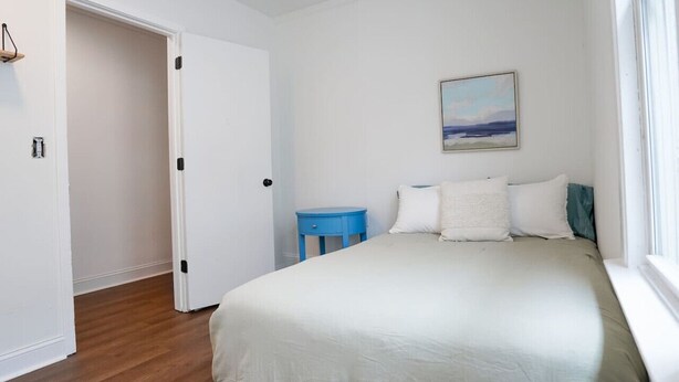 Une chambre à coucher avec un lit, une table de chevet bleue, un tableau accroché au mur et une porte donnant sur une autre pièce.
