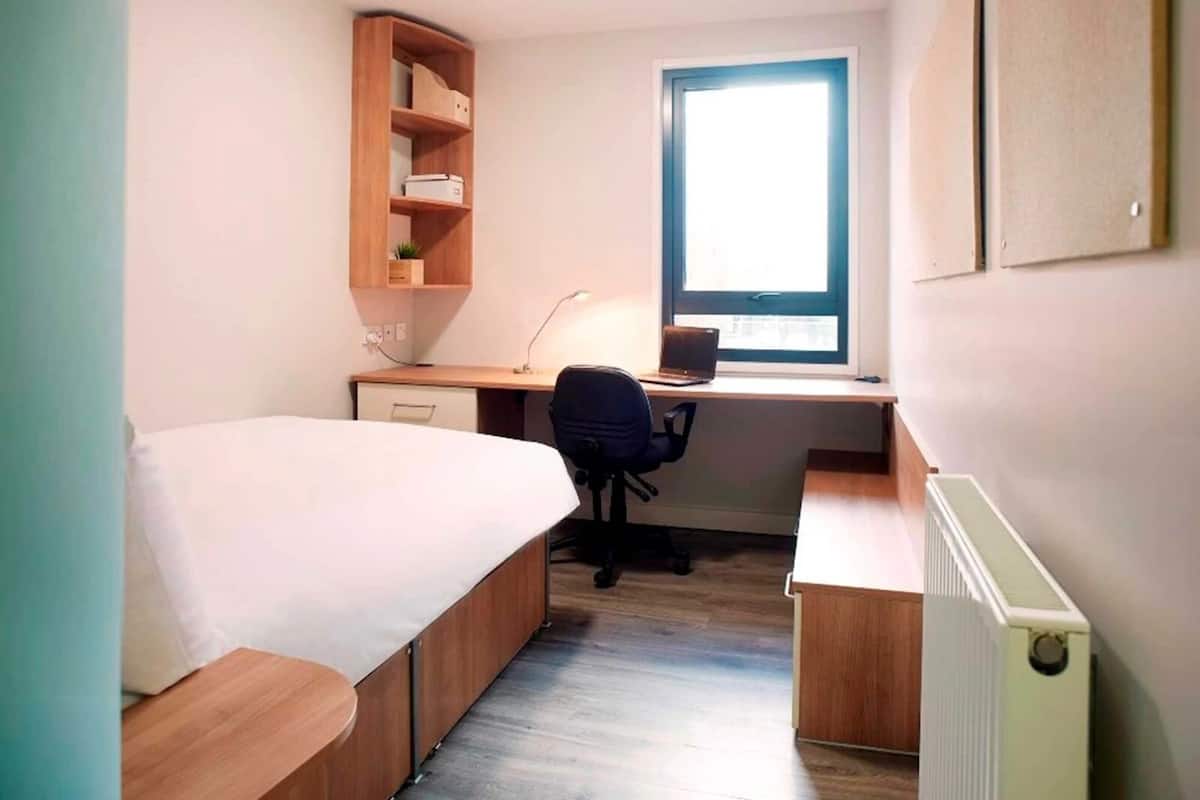 A small, well-lit room with a bed, a desk with a chair, a window, and a radiator.