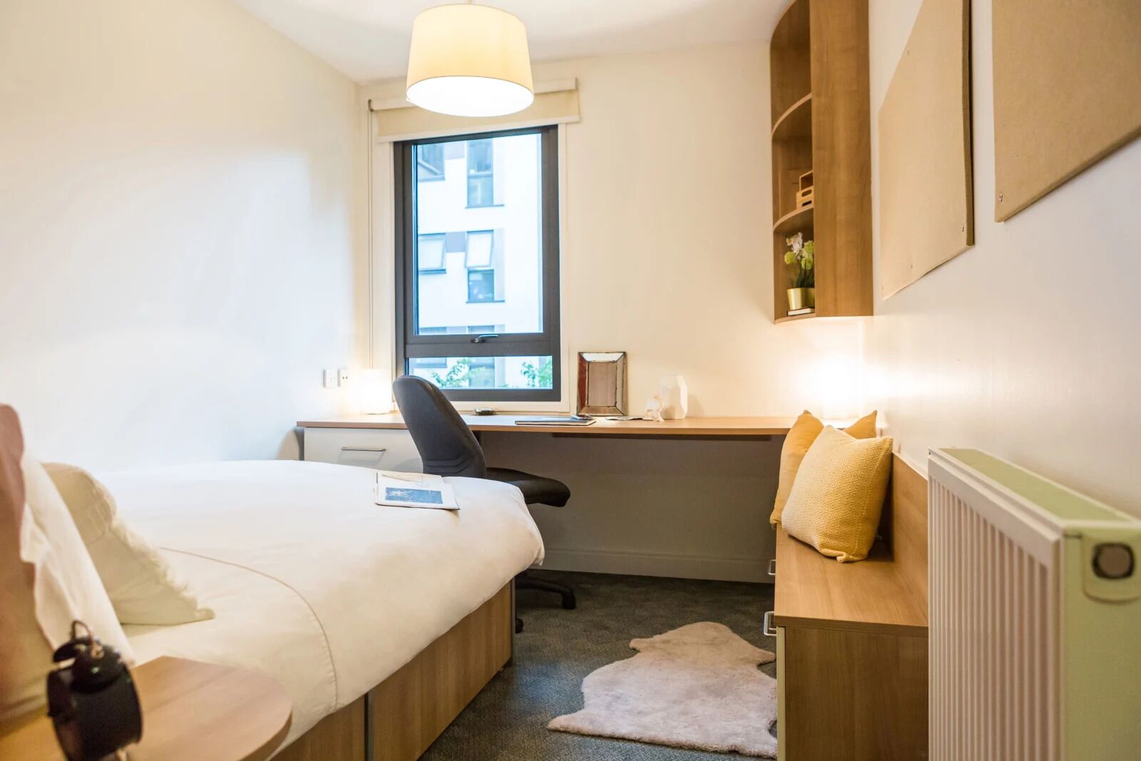 A compact hotel room with a bed, a desk, a chair, a radiator, and a window with a view of buildings.