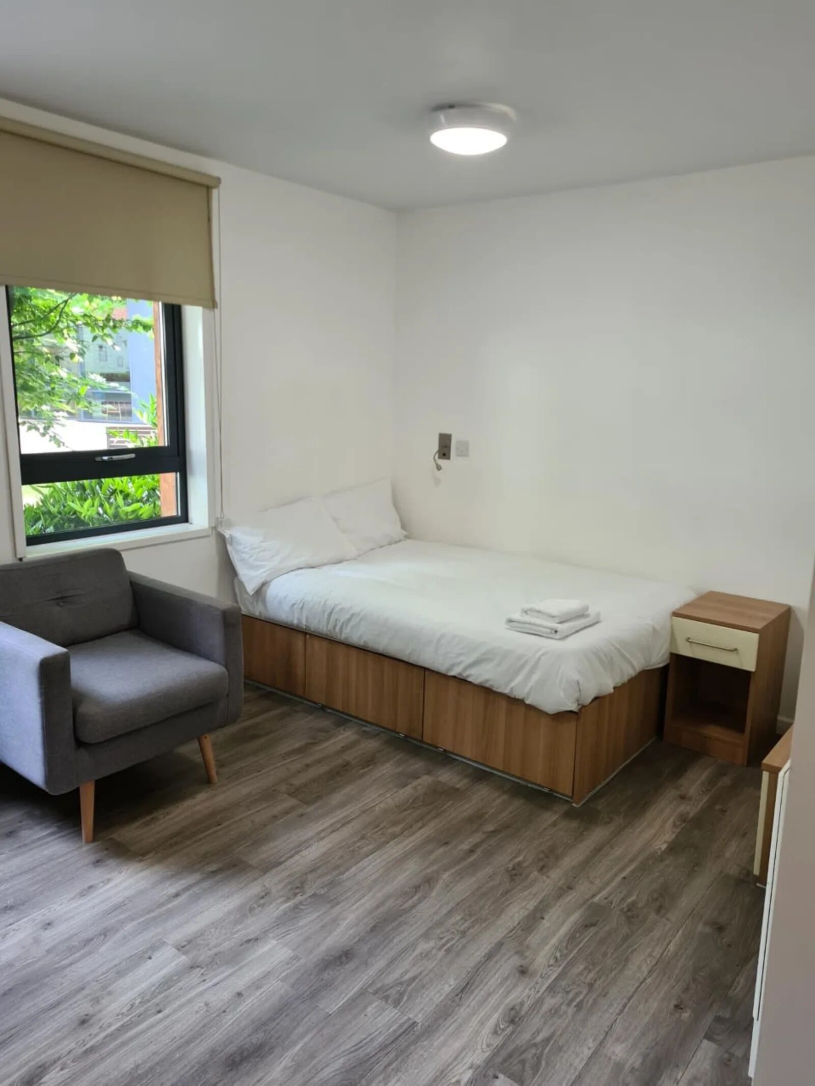 A small, clean room with a bed, a chair, a nightstand, and a window with a view of greenery outside.