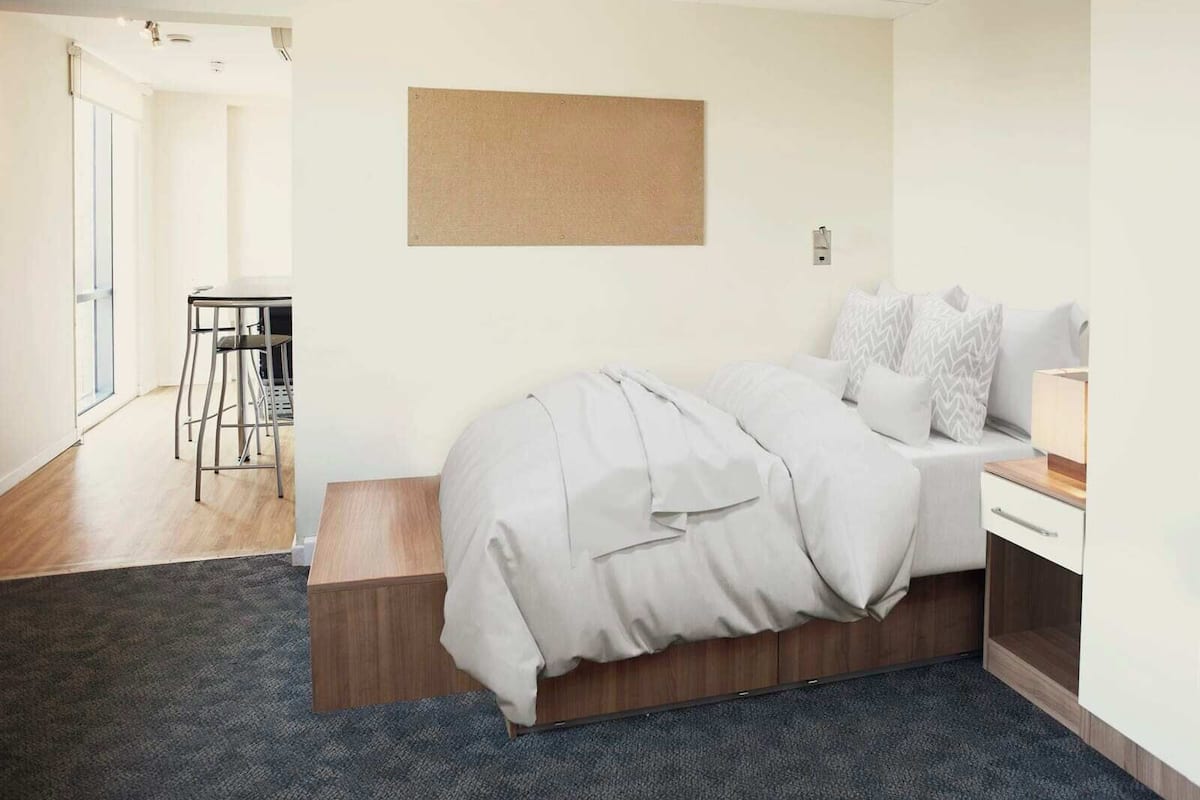 A modern bedroom with a bed, bedside table, and a desk. There is a window and a door leading to another room.