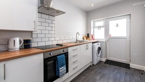 A modern kitchen with white cabinets, a wooden countertop, a kettle, a coffee mug, a stove, a washing machine, and a door with an anchor decoration. - Maritime House II (Hull)