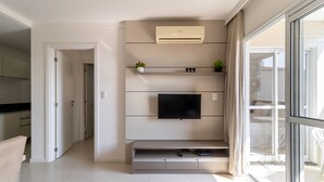 A modern living room with a flat-screen TV mounted on a wall, a built-in air conditioning unit above it, and a sliding glass door leading to an outdoor area. - IAPs Apto 140m da praia Floripa (Florianópolis)