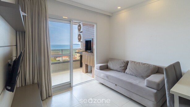 A modern living room with a sofa, a TV, and a sliding glass door leading to a balcony with a view of the sea. - IAPs Apto 140m da praia Floripa (Florianópolis)
