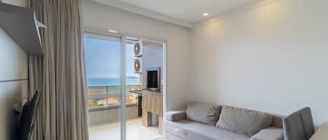 A modern living room with a sofa, a TV, and a sliding glass door leading to a balcony with a view of the sea.