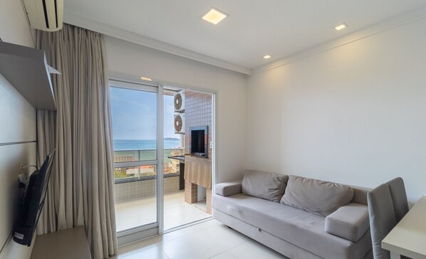Un salón moderno con sofá, televisor y puerta corrediza de vidrio que da a un balcón con vistas al mar.