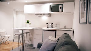 A compact kitchen with a microwave, a small refrigerator, and a sink. There is a dining area with a white table and two bar stools. A grey sofa with a cushion is positioned in front of the kitchen. - Italianway - Copernico I (Milan)