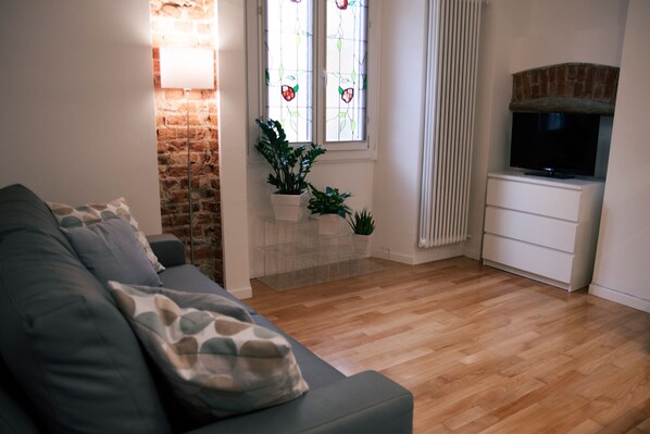 A living room with a grey sofa, wooden flooring, a TV on a white cabinet, a brick fireplace, and a window with stained glass.
