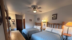 Ein Schlafzimmer mit zwei Betten, einer hölzernen Bank, einem Deckenventilator, einem Nachttisch mit Lampe und einem durch eine offene Tür einsehbaren Badezimmer.