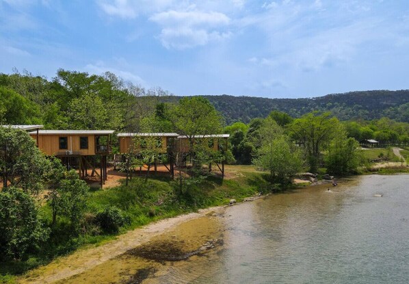 A riverside property with wooden cabins elevated on stilts, surrounded by lush greenery and a clear sky. - Waterfront Sky Cabin 2 by Best Texas Travel (New Braunfels)