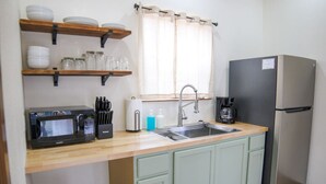 A compact kitchen with a microwave, sink, and refrigerator. There are shelves above the counter holding dishes and jars.