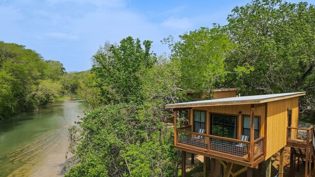 A wooden treehouse with a balcony, situated by a river surrounded by dense greenery.