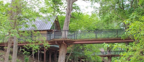 A rustic cabin with a metal bridge connecting two levels, surrounded by dense forest.