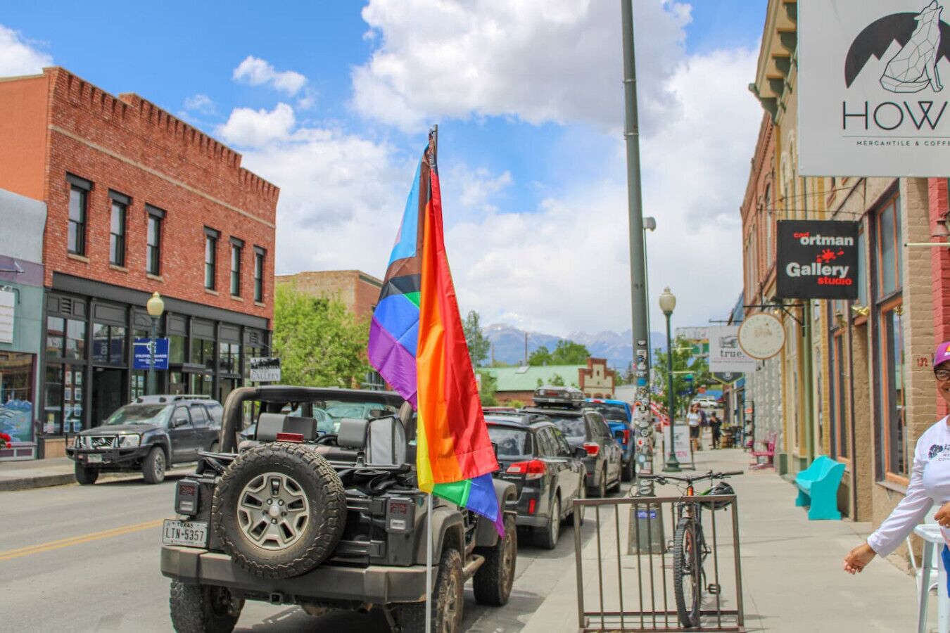 A street with a rainbow flag, a Jeep, and a storefront with a sign that reads 'HOW'.