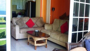Un salón con un sofá beige, cojines rojos, una mesa de centro de mimbre y vistas a la cocina y al exterior.