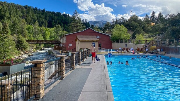 Ein Pool mit einem gepflasterten Weg, ein rotes Gebäude und ein bewaldeter Hang im Hintergrund.