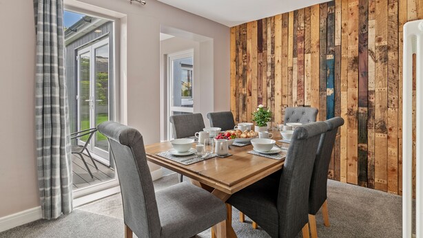 A dining area with a wooden table set for six, grey upholstered chairs, and a view through a glass door to a balcony. - Roslen - Lovely Bungalow Near Beach and Village (Saundersfoot)